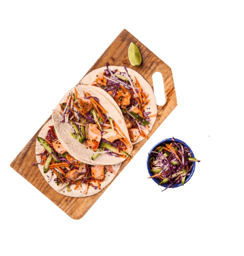 Three salmon tacos filled with shredded vegetables served on a wooden board with a lime wedge