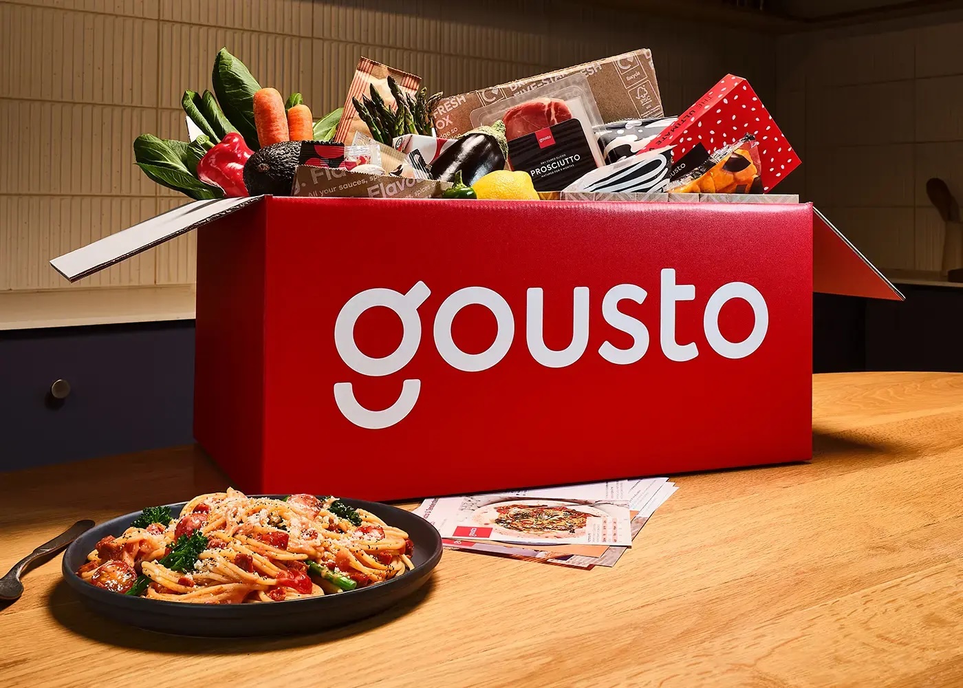 Open Gousto recipe box containing vegetables, meat, spices, and recipe cards, next to a bowl of creamy tomato sauce pasta cooked recipe.