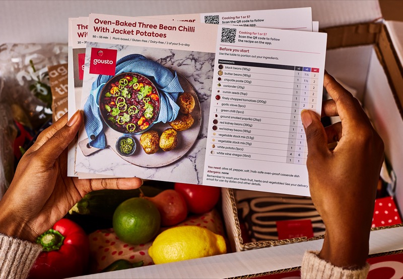 Customer holds recipe cards above pre-portioned ingredients inside an insulated Gousto box.