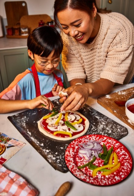 Parent and child making a homemade pizza together in the kitchen, adding toppings to the dough