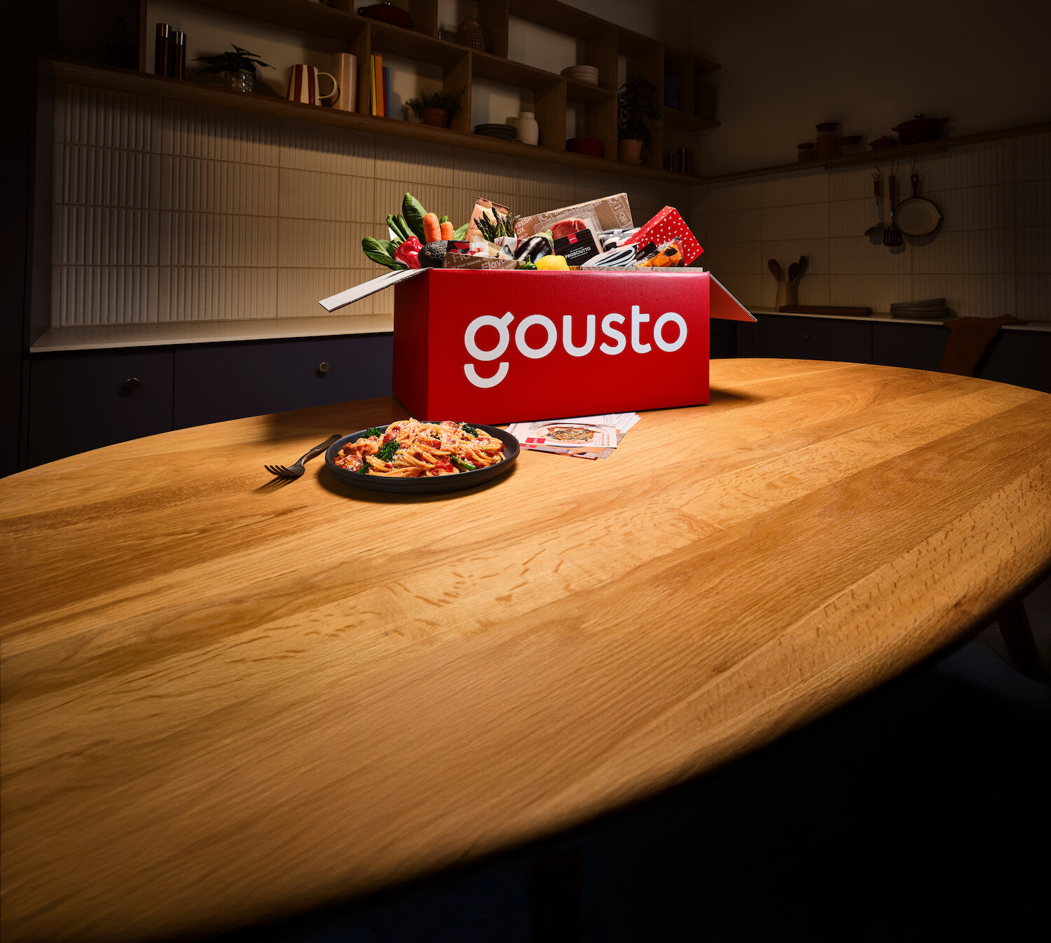 A kitchen counter with a Gousto box overflowing with fresh ingredients and a plated meal