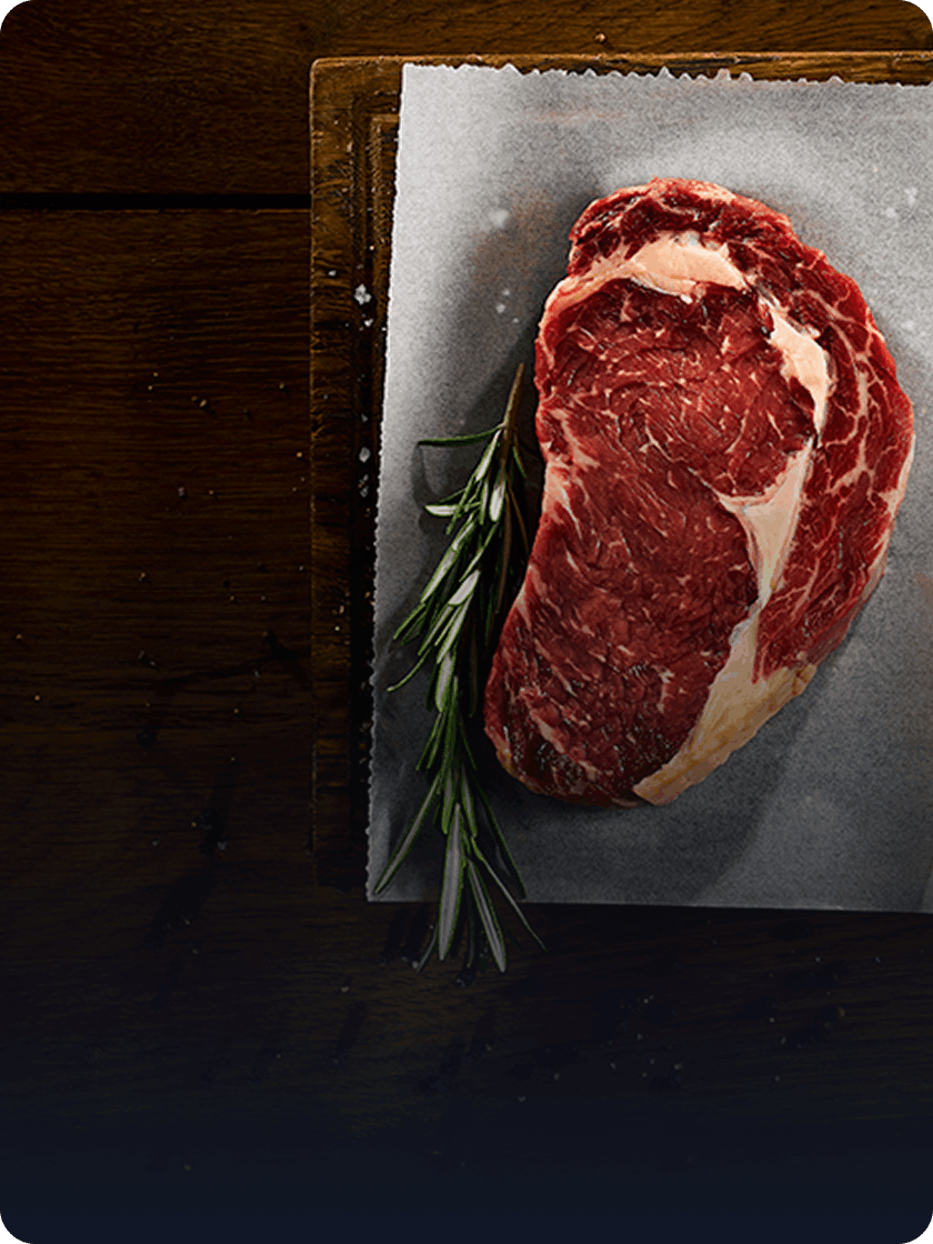 Overhead shot of a raw steak with rosemary, a meat cleaver and peppercorns on a wooden board