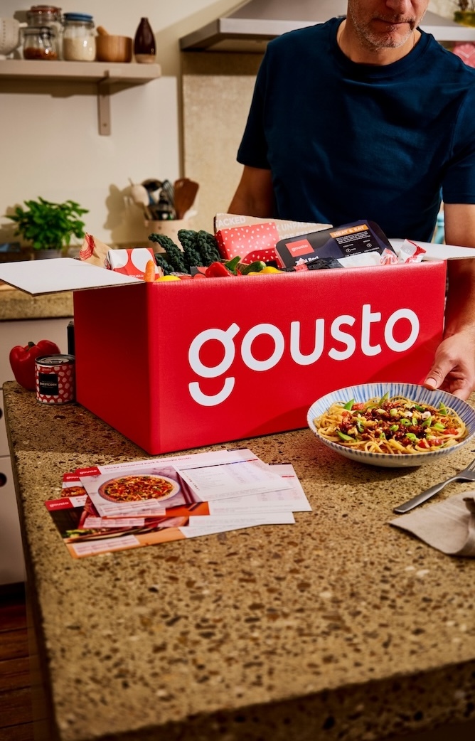 A man in a home kitchen unpacking a Gousto box of fresh ingredients on the counter, next to a bowl of cooked noodles and a recipe card