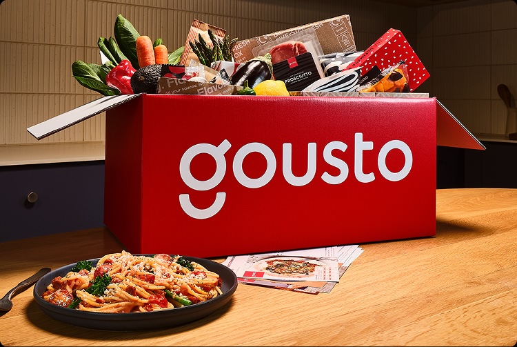 Gousto recipe box containing vegetables, meat, spices, and recipe cards, next to a creamy tomato sauce pasta cooked recipe.