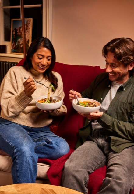 Couple sitting on a red sofa smiling and eating noodle bowls together for a date night at home