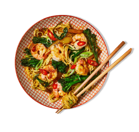 A dish of prawn noodles with fresh broccoli, red chili slices, and spring onions, served in a patterned bowl
